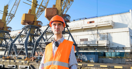 male worker of sea harbor in orange helmet and safety west, cranes and sea background 