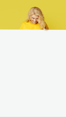 Wall Mural - Young happy woman peeks out from behind a white banner on a yellow background. Point to an empty blank on a form, a copy space for text. vertical shot