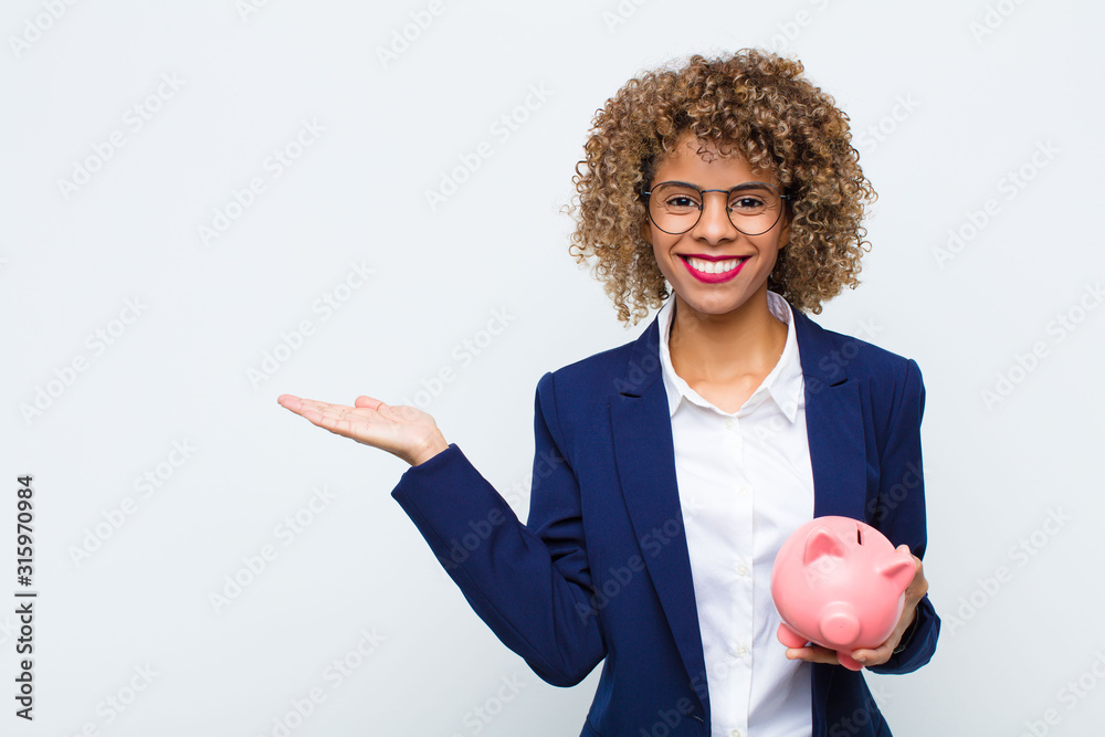 young african american woman smiling cheerfully, feeling happy and showing a concept in copy space with palm of hand with a piggy bank - obrazy, fototapety, plakaty 