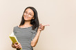 © Asier - Young asian woman student holding a book smiling and pointing aside, showing something at blank space.