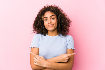 Wall Mural - Young african american woman against a pink background points sideways, is trying to choose between two options.