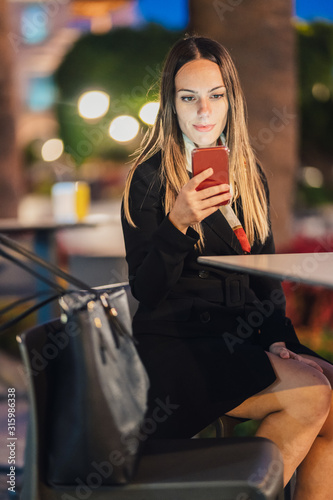 Fotomural business woman sitting on a terrace at night checking her mobile phone