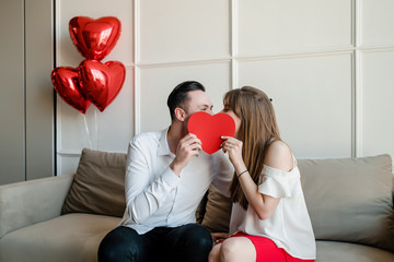 man and woman with red heart shaped valentine card and balloons at home on couch