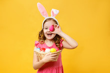Happy Child In The Ears Of A Bunny Holds An Easter Egg And A Basket. Portrait Of A Little Girl On A Yellow Background