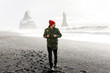 young male traveler in green clothes in a red hat and a green backpack goes black beach in Iceland