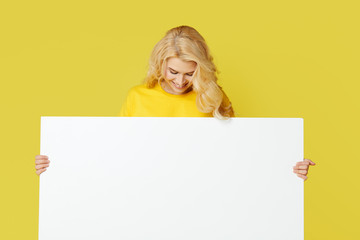 Wall Mural - Young happy woman peeks out from behind a white banner on a yellow background. Point to an empty blank on a form, a copy space for text. Horizontal shot