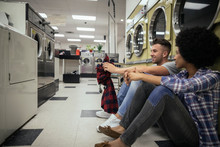 Young Couple Sitting And Talking While Waiting For Laundry At Laundromat