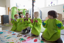 Playful Preschool Students In Smocks Showing Finger Paint On Hands At Poster In Classroom