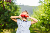 Fototapeta Młodzieżowe - Adorable little female picked two fresh ripe apples from apple tree and holds them infront of her eyes, grinning to the camera.