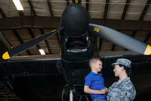 Female Army Pilot Mother Holding Son Below Airplane In Airplane Hangar