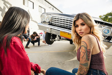 Portrait Confident Latinx Young Woman With Tattoos In Front Of Low Rider Car Bouncing In Parking Lot