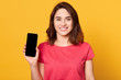© sementsova321 - Horizontalshot of young woman showing her mobile cell phone with black screen over yellow background, adorable female wearing casual t shirt, looking smiling directlyt at camera. Copy space.
