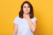 Close up portrait of cute sick young brunette woman wearing white casual t shirt, having sore throat, holding hand on her neck, sufferingfrom throat pain, painful swallowing, health care concept.