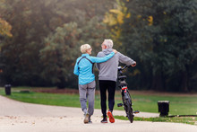 Cheerful Active Senior Couple With Bicycle Walking Through Park Together. Perfect Activities For Elderly People.