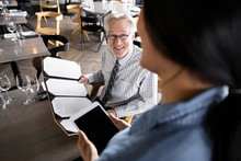 Waitress With Digital Tablet Taking Order From Smiling Senior Businessman At Restaurant Table
