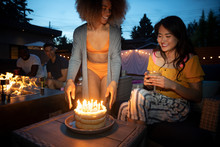 Happy Young Woman Serving Birthday Cake To Friend On Summer Patio At Night