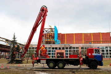 Firefighter rescuers at work in fireproof suits came to extinguish a fire in a fire truck and pull out a ladder cradle lift at a large industrial plant with pipes and equipment.