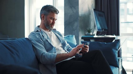 Wall Mural - Handsome caucasian mature man sitting on the sofa at home and using phone watching video or contents serfing internet technology modern apartment social media browsing isolated
