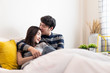 © Kawee - Lovely Asian couple sitting on bed in bedroom together. Boy holding girl head softly and be hand in hand or pinky swear to love girl. Man kissing woman on smile young lady cheek feeling shy & happy.