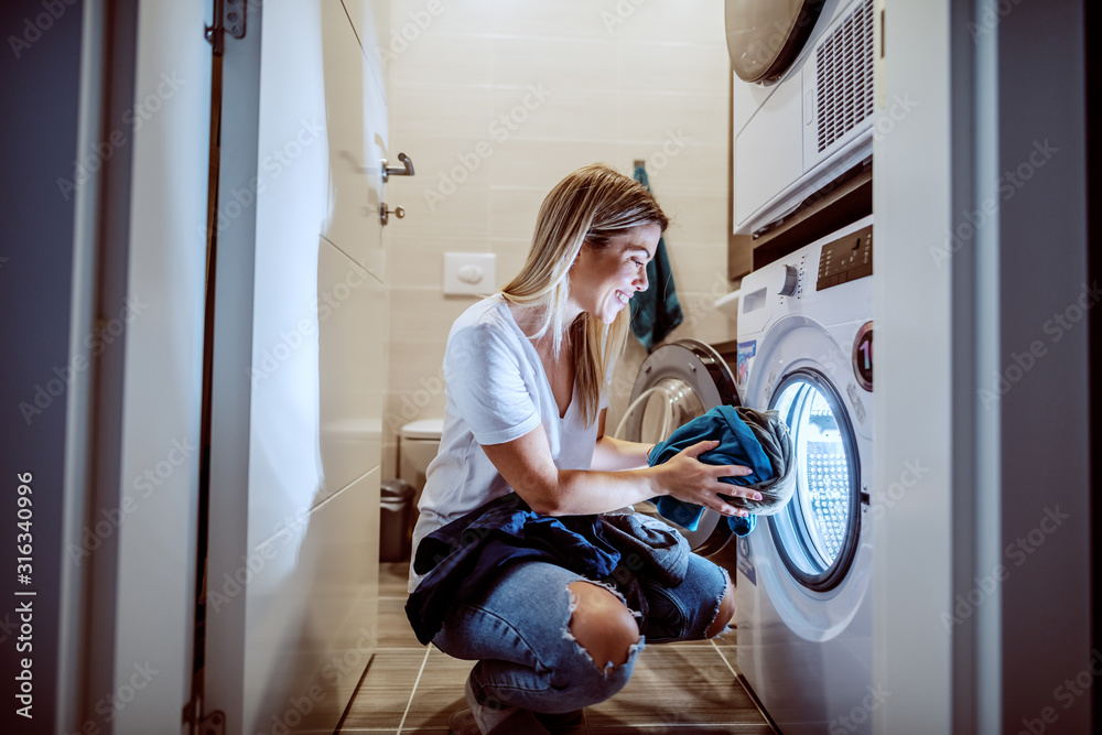 Smiling caucasian blond worthy housewife dressed casual crouching in bathroom and loading washing machine with dirty clothes in late hours. - obrazy, fototapety, plakaty 