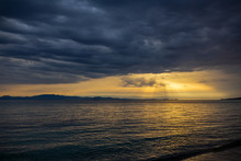 Horizontal Color Photography Of Amazing Dramatic Sunset Or Sunsrise Landscape Of Dark Heavy Grey Clouds, Transparenting Through Them Soft Sun Beams Falling Dawn On Dark Surface Of Sea Water. Greece. 