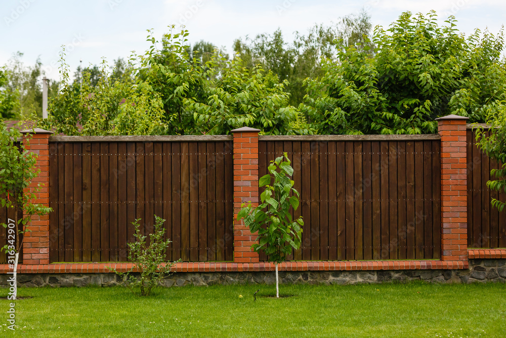 New wooden fence with massive stone brick pillars. Green lawn and trees, daytime
