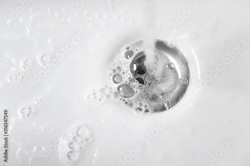 Fotografía Water with soap foam in white ceramic sink drains down, close up