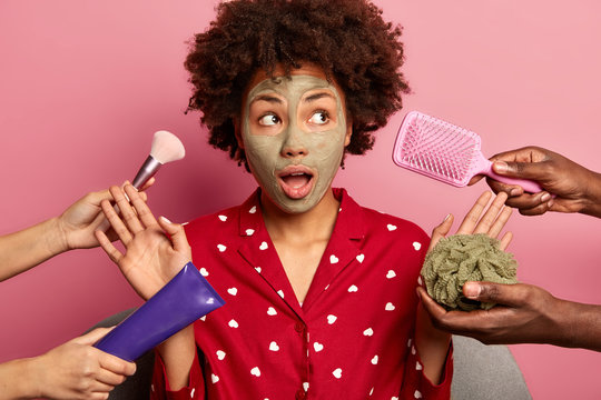 Surprised beautiful woman wears red night clothes, applies clay mask on face, raises palm towards various beauty products, shocked by so much suggestions at same time, looks away with opened mouth