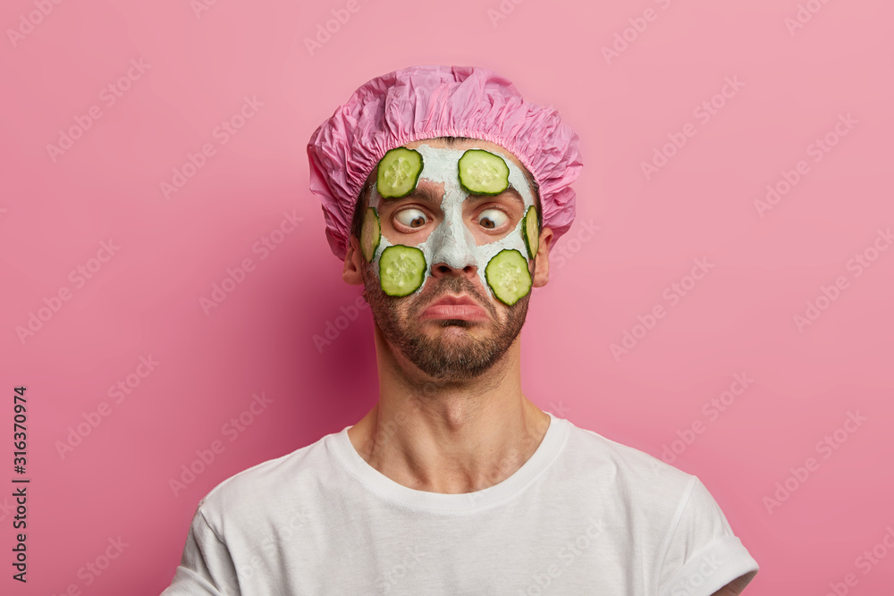 Young man crosses eyes, applies cream mask on face with cucumber, has beauty session, wears bath cap, reduces dark dotes on complexion, isolated over pink background. Facial treatment concept - obrazy, fototapety, plakaty 