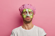 © wayhome.studio  - Young man crosses eyes, applies cream mask on face with cucumber, has beauty session, wears bath cap, reduces dark dotes on complexion, isolated over pink background. Facial treatment concept