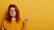 Photo of lovely ginger woman points index finger aside, demonstrates promo on right, looks with interesting expression, has wavy red hair, presents promo, recommends advertisement over yellow wall.