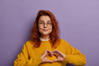 © WHstudio Leushin N - Romantic redhead young woman shows heart gesture over chest, smiles pleasantly, wears transparent glasses and yellow jumper, poses during photoshoot against purple background. Love confession