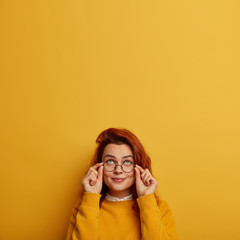 Wall Mural - Indoor image of thoughtful redhead Caucasian woman keeps hands on frame of glasses, tries to see something above, has curious glad expression, wears yellow jumper. People, daydreaming concept