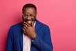 © wayhome.studio  - Photo of overjoyed Afro American man laughs at funny story, cant stop giggling, has white teeth, thick beard, wears formal suit, squints face, isolated on pink background, glad to achieve success