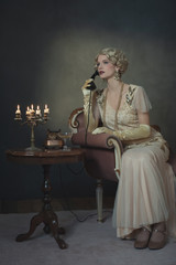 Retro 1920s fashion woman on the phone while sitting on sofa beside table with candlestick and glass of champagne.