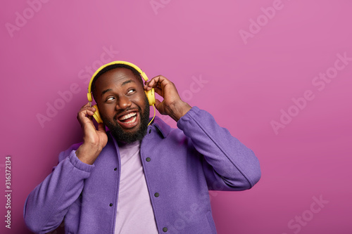 Fotografía Horizontal view of positive guy wears stereo headphones on ears, enjoys live music, looks aside, being music meloman, dressed casually, has hobby, isolated over vivid studio wall