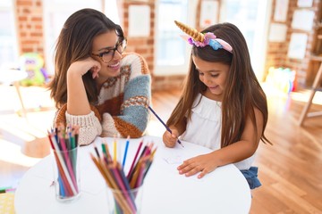 Wall Mural - Beautiful teacher and toddler wearing unicorn diadem drawing using paper and pencils around lots of toys at kindergarten