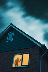 Poster - Intruder with handgun in eerie house with illuminated window under stormy sky at dusk.