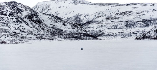 Wall Mural - two people crossing a frozen lake on foot surrounded by rocky mountains in a polar environment north