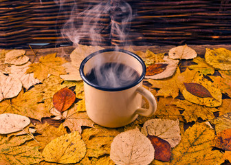 Wall Mural - Hot coffee with steam in an enamel mug on a background of leaves. Autumn cozy background.