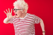 © Krakenimages.com - Young albino blond man wearing striped t-shirt and glasses over isolated red background shouting and screaming loud to side with hand on mouth. Communication concept.