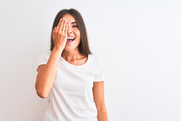 Wall Mural - Young beautiful woman wearing casual t-shirt standing over isolated white background covering one eye with hand, confident smile on face and surprise emotion.
