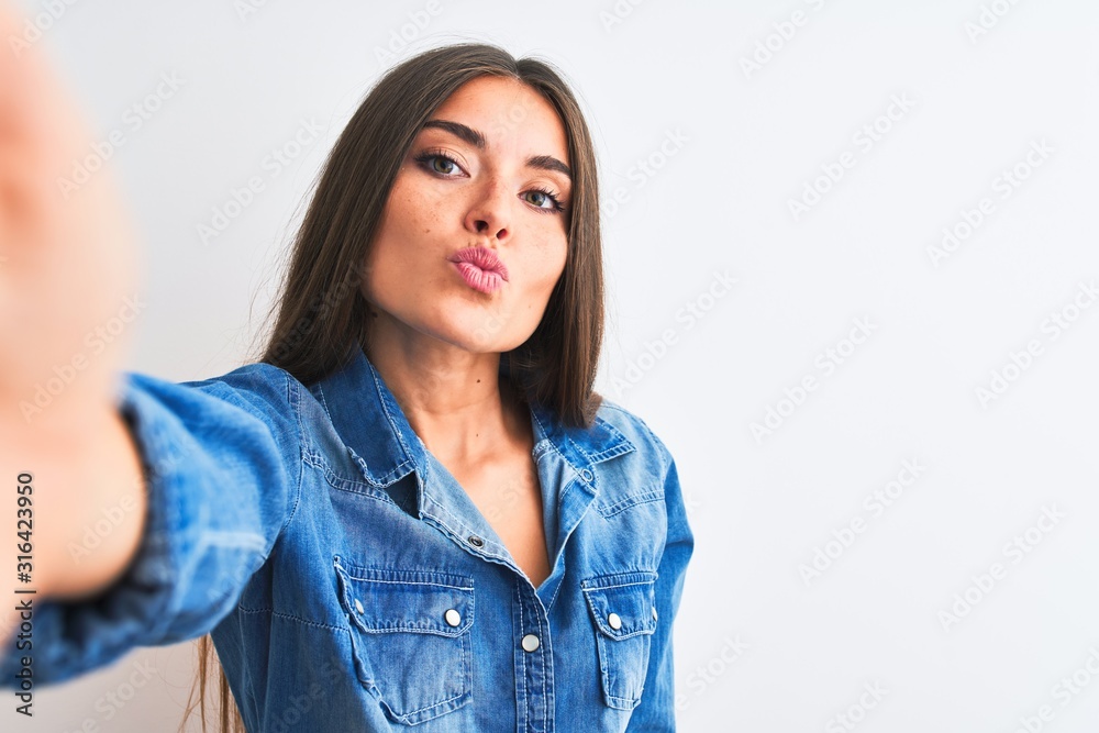 Beautiful woman wearing denim shirt make selfie by camera over isolated white background looking at the camera blowing a kiss on air being lovely and sexy. Love expression. - obrazy, fototapety, plakaty 