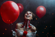 Close-up Portrait Female Clown Woman Scary Crazy Smiling Laugh Shows Sharp Teeth Predator Face, Holding Red Balloons. Backdrop Dark Gothic Room Sparks Fire Hell. Art Creative Bright Halloween Make-up