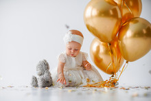 A One Year Old, Baby Girl Sitting With A Bunch Of Golden Balloons And Taddy Bear On White Background In Studio.