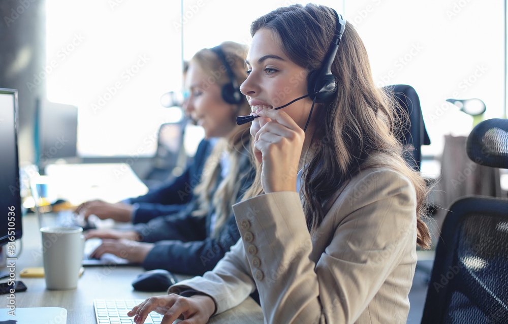 Female customer support operator with headset and smiling, with collegues at background. - obrazy, fototapety, plakaty 