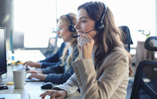 Female Customer Support Operator With Headset And Smiling, With Collegues At Background.