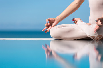 Side view of a unrecognizable young slender woman sitting in lotus position and meditating on the edge of the pool on a blue sky. Concept of yoga relaxation and meditation. Copyspace. Mudra