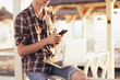 © Rithor - Positive young guy a freelancer works with a laptop and chats with friends using a smartphone on a sunny summer day on a beach under a thatched canopy