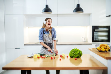 Young happy caucasian woman in apron standing in kitchen and cutting cucumber while listening music and singing. On kitchen counter are all sorts of vegetables. Healthy eating concept.